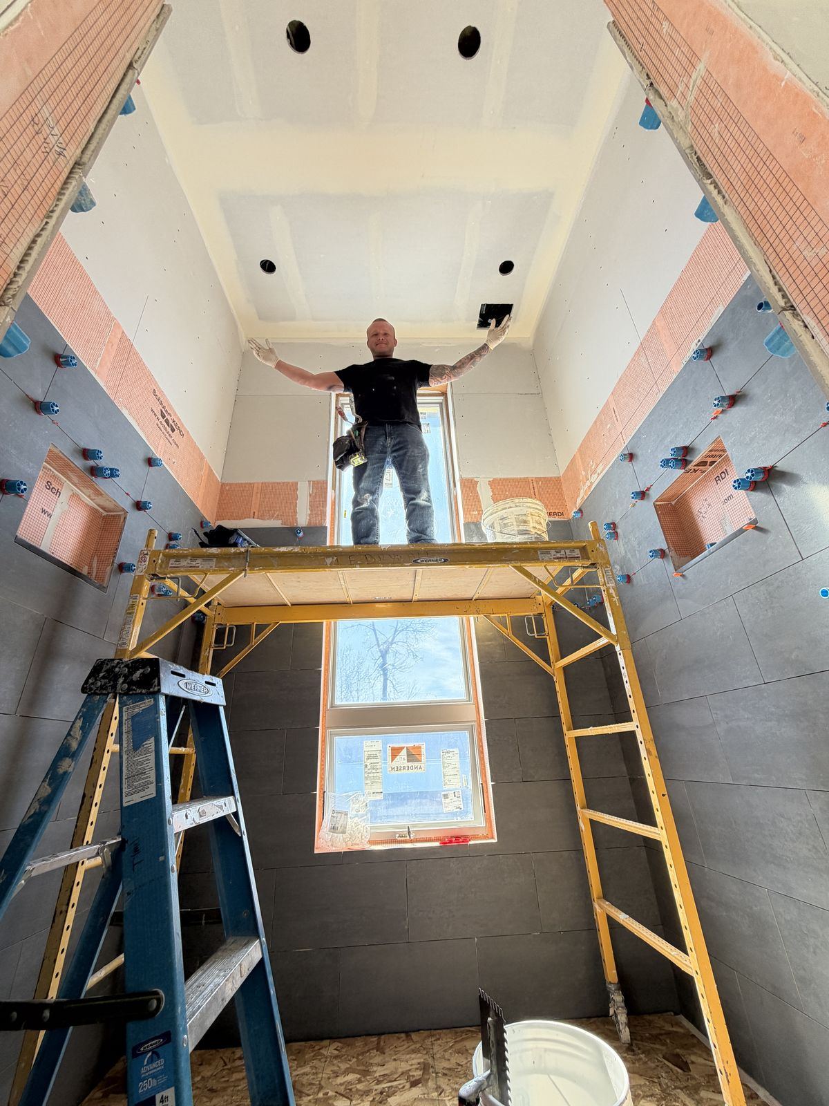 Micah on a scaffold in a tall shower, waterproofing wrapped floor to ceiling, large-format tile going up
