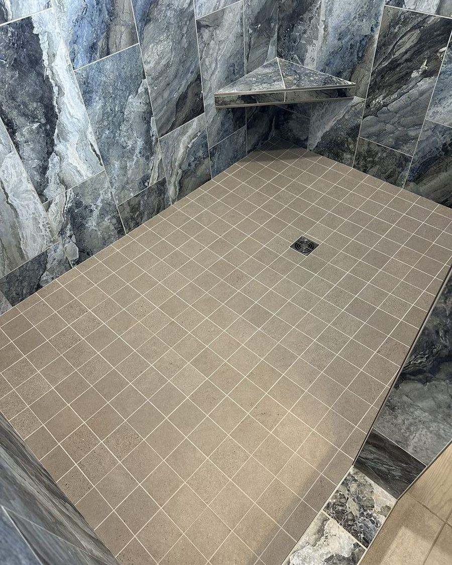 Finished shower floor: small mosaic tile sloped to a square drain, with marble-look tile walls and a corner shelf